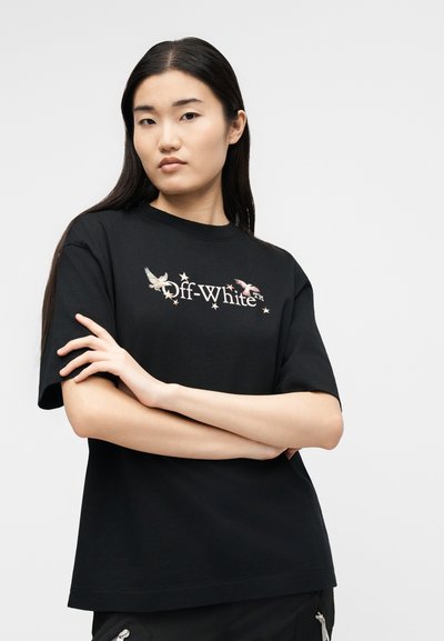 Γυναίκα με μακριά σκούρα μαλλιά φοράει μαύρο t-shirt Off-White με γραφικά πουλιού και αστέρι, με σταυρωμένα χέρια μπροστά σε απλό φωτεινό φόντο.