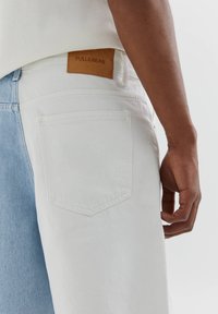 Pantalon en denim blanc et bleu clair avec une étiquette Pull&Bear en cuir marron, doté d'une poche arrière et d'une coupe décontractée.