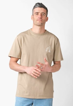 Rövid, só-bors színű hajjal rendelkező férfi, bézs színű pólóban, melyen a mellrészen kis fehér szörfdeszka grafika látható, világoskék farmert visel, kezeit összekulcsolva.