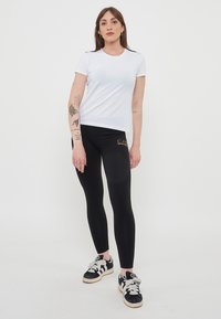 T-shirt bianco a maniche corte abbinato a leggings neri con logo del marchio dorato. Modella che indossa sneakers nere con strisce bianche e lacci.