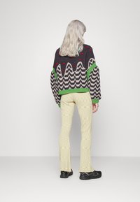 Strikket sweater med et zigzag mønster i grå, grønne accenter og røde prikker; parret med teksturerede lysegule bukser og sorte sko.