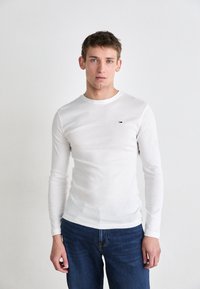 Vit långärmad skjorta i ett lätt material, rund halsringning, figurnära design, med en liten logotyp på bröstet. Kombinerad med blå jeans.