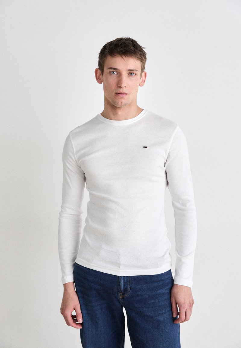 Vit långärmad skjorta i ett lätt material, rund halsringning, figurnära design, med en liten logotyp på bröstet. Kombinerad med blå jeans.