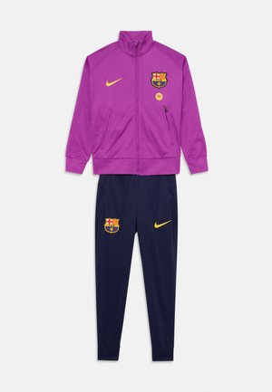 Survêtement du FC Barcelone comprenant une veste zippée violette et un pantalon bleu marine. Matériau : polyester. Les accents incluent le logo de l'équipe et le swoosh de Nike en jaune.