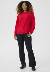 Punainen neuletakki, jossa on venepääntie, linnunselkähihoja ja ribbireunat, yhdistetty tummansiniset leveälahkeiset farkut ja mustat kiiltävät kengät.