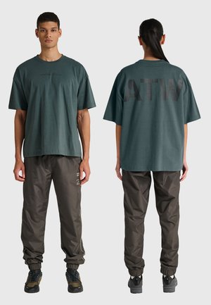 Man in een donker groen oversized T-shirt met "ATW" op de achterkant en bruine joggerbroek, van voren en van achteren getoond op een neutrale achtergrond.