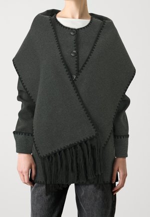 Mørkegråt uldponcho med sort zigzagkant, sorte kvaster ved kanten, båret over en hvid top og mørke bukser, vist på en person fra nakke til hofter.