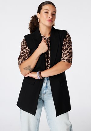Femme aux cheveux bouclés en queue de cheval porte un haut imprimé léopard, un blazer noir sans manches, un jean bleu clair et des bracelets violets et marron.