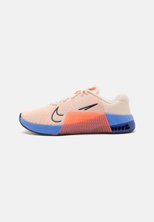 Zapatilla deportiva de malla beige con logo negro, entresuela naranja y suela azul. Presenta un diseño texturizado y un sistema de cordones para un ajuste seguro.