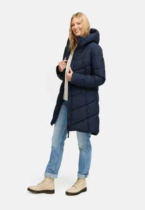 Marineblaue Daunenjacke mit Kapuze, diagonal gesteppt und mit Reißverschluss. Kombiniert mit hellblauen hochgekrempelten Jeans und beigen Schnürstiefeln.