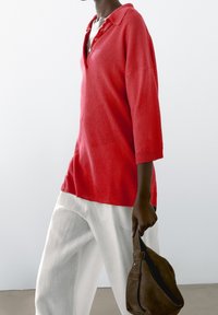 Czerwony dziergany sweter polo z kołnierzykiem, rękawami do połowy przedramienia i prążkowanym wykończeniem, zestawiony z luźnymi białymi lnianymi spodniami oraz ciemnobrązową torbą.