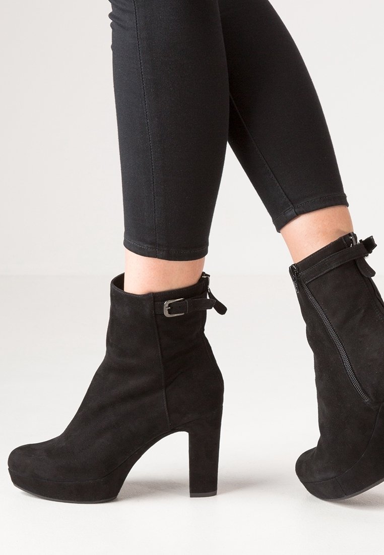 Bottines en daim noir avec un talon bloc haut, une fermeture éclair latérale et un détail de boucle ajustable sur le côté extérieur. Texture lisse dans l'ensemble.