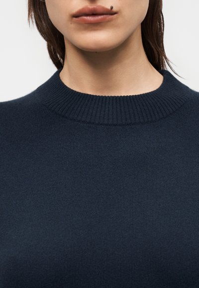 Gros plan d'une personne portant un pull côtelé à col rond bleu marine foncé, montrant le bas du visage, le cou et le haut de la poitrine.