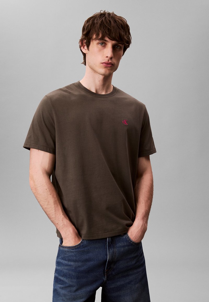 Jeune homme aux cheveux bruns portant un t-shirt à manches courtes vert olive avec un logo rouge "cK" et un jean bleu, les mains dans les poches, sur un fond gris uni.