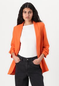 Orange skræddersyet blazer med spidse revers, oprullede ærmer og en afslappet pasform, båret over en hvid top og mørke jeans. Glat tekstur.