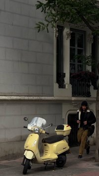 Vespa skuter v pastelno rumeni barvi z jasno vetrobransko steklo, črnim sedežem in zadnjo prtljažno omaro, parkiran poleg stavbe z okrašenim zasnovam.