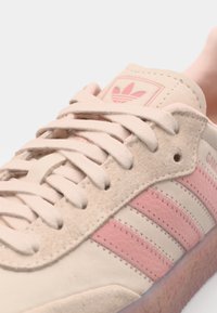 Beige Wildledersneaker mit pinken Lederstreifen, runden Schnürsenkeln und einem Logopatch auf der Zunge; strukturierte Gummisohle.