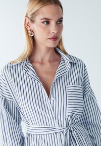 Valge triiksärk tumesiniste vertikaalsete triipudega, millel on rinnahoidja tasku ja nööritav vöökoht. Sile kangas, krae ja pikad varrukad.