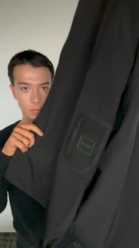 Νεαρό άτομο κρατά μαύρο μπουφάν με ορατή τσέπη με φερμουάρ και ετικέτα "BOSS" μπροστά σε απλό φόντο.