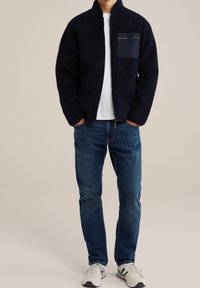 Fleece zip-up jacka i marinblått med en strukturerad yta, med en bröstficka i matchande material. Bärs tillsammans med blå jeans och sneakers.