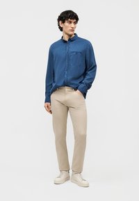 Mann iført blå skjorte med knapper, beige bukser og hvite joggesko, står med en hånd i lommen mot en ensfarget bakgrunn.