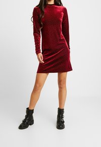 Röd sammetsklänning med långa ärmar och vita prickar, figurnära design, midjehöjd, kombinerad med svarta ankelstövlar.