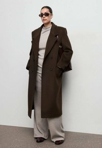 Manteau croisé marron foncé, pull gris et pantalons larges, lunettes de soleil, sac à épaule, textures lisses, design minimaliste.