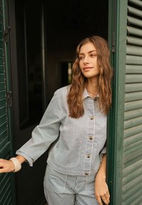 Femme aux longs cheveux ondulés portant une veste rayée boutonnée et un pantalon, se tenant dans une porte, tenant des volets verts ouverts, regardant vers la droite.