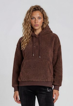 Jeune femme aux cheveux blonds ondulés portant un sweat à capuche en peluche marron et un pantalon noir sur un fond gris uni, regardant vers l'avant.