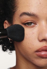Pinceau de maquillage noir appliquant un produit sur un visage texturé, légèrement tacheté de rousseur, avec des lèvres pulpeuses. Les tons de maquillage naturels rehaussent l'apparence de la peau.