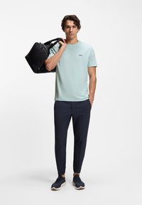 Άνδρας που φοράει ανοιχτό μπλε μπλουζάκι με το λογότυπο "BOSS" και σκούρο παντελόνι κρατάει μαύρη τσάντα duffel στον ώμο μπροστά από λευκό φόντο.