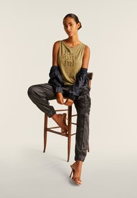 Canotta verde oliva con stampa metallica, abbinata a pantaloni jogger neri in satin e a una giacca blu navy, su uno sfondo neutro.