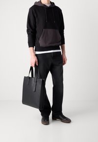 Bolso tote de cuero negro con forma plana, asas cortas y diseño minimalista. Lleva puesto con una sudadera negra y jeans oscuros.