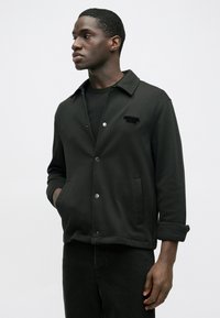 Mustast puuvillast jope kraega kaelusega, trukkidega ja küljetaskutega. Rinnal reljeefse tekstilogoga ning üleskeeratud varrukatega.