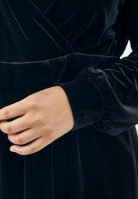 Melnā samta kleita ar iesienamu dizainu un savilkta piedurkņu galiem, ar gludu tekstūru un pielāgotu jostasvietu, uzsverot elegantu siluetu.