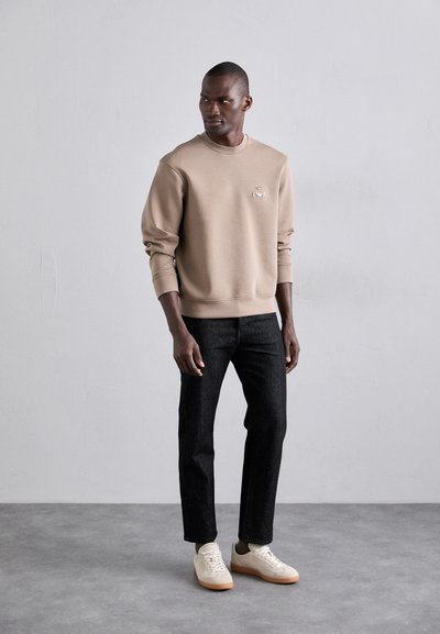 Beige sweatshirt med rund hals og brodert logo, kombinert med mørke denimjeans og hvite joggesko, stående på en nøytral bakgrunn.