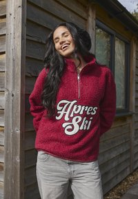 Czerwony polar z zamkiem w kołnierzu, z napisem "Apres Ski" w białym kursywie. Połączony z jasnoszarymi dżinsami na tle drewnianym.