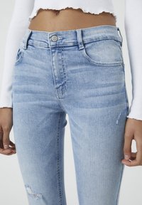 Jean en denim bleu clair au design ajusté, avec une taille mi-haute, un style à cinq poches et de légers détails déchirés sur la cuisse.