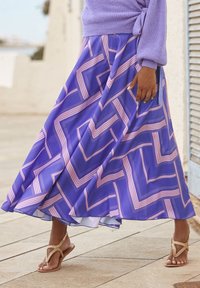 Długa fioletowa spódnica w wzór chevron w różowe i białe paski, zwiewny materiał, połączona ze złotymi sandałami na paski. Miękka tekstura, długość do połowy łydki.