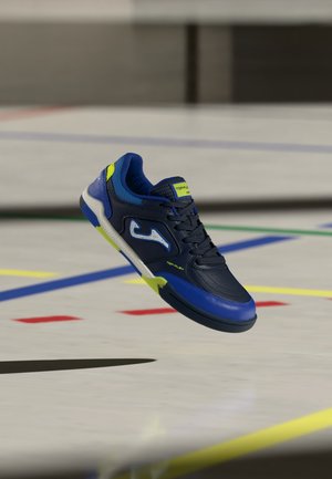 Granatowo-jaskrawoniebieski but sportowy do użytku wewnętrznego z żółtymi akcentami unoszący się nad podłogą sali gimnastycznej oznaczoną kolorowymi liniami.