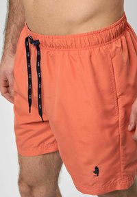 Korálové plavecké šortky vyrobené z ľahkého materiálu, s elastickým pásom, námorníckym šnúrkou na sťahovanie a malým čiernym vyšitým logom.