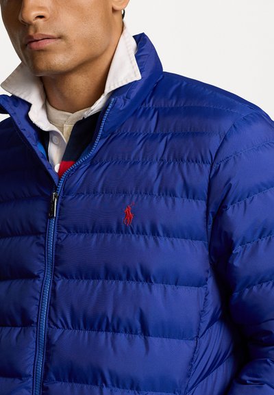 Casaco puffer azul com um design acolchoado, logótipo vermelho em contraste no peito e gola alta. Fecho com zipper. Tecido suave e brilhante.