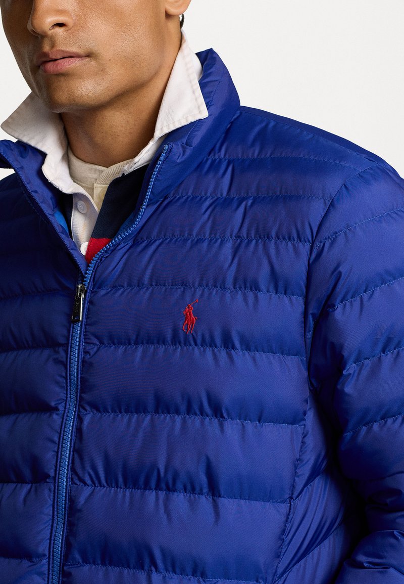 Blauer Steppjacke mit einem gesteppten Design, kontrastierendem rotem Logo auf der Brust und einem Stehkragen. Reißverschlussverschluss. Glattes, glänzendes Material.