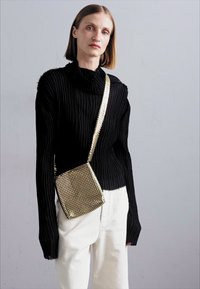 Un pull à col roulé noir côtelé associé à un pantalon blanc. Un sac bandoulière en maille métallique dorée est porté en travers du corps, ajoutant un contraste.