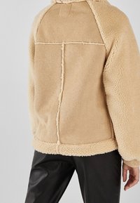 Tylny widok osoby w beżowej kurtce z owczej skóry z widocznymi szwami i czarnych skórzanych spodniach na jednolitym tle.