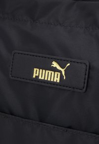 Czarna faktura materiału z prostokątną czarną metką z wytłoczonym złotym napisem "PUMA" oraz złotym logo skaczącego pumy.