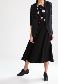 Svart kavaj över en blommig blus, kombinerad med en svart midiklänning och svarta sneakers. Slät tyg, figurnära design och avslappnad stil.