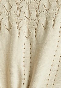 Tissu en tricot crème avec motifs symétriques en dentelle diamant et motifs œillets verticaux, montrant la texture et les détails du travail de couture.