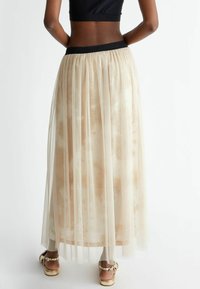 Lunga gonna plissettata beige realizzata in tulle trasparente, caratterizzata da una vita elasticizzata nera con dettaglio del logo. La finitura metallica aggiunge una texture sottile.