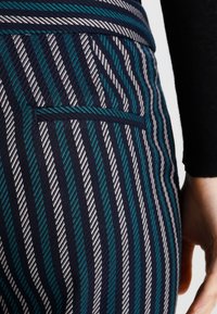 Randiga byxor i marinblått, vitt och teal-tyg. Slim fit med en bakfickdetalj, som visar ett strukturerat mönster.